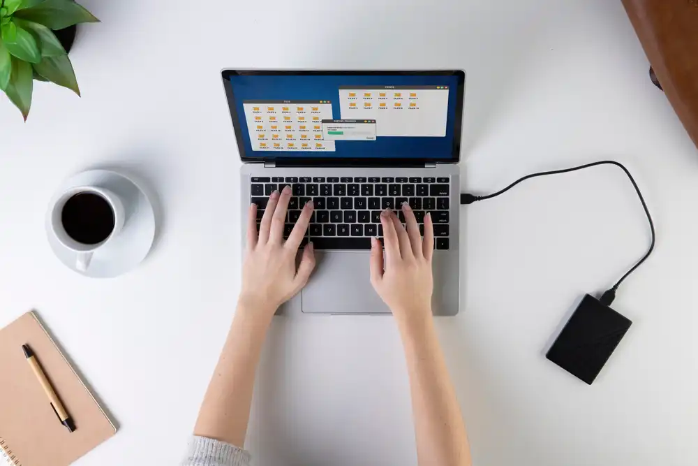 لپ تاپ متصل به هارد اکسترنال در کنار فنجان قهوه و دفترچه یادداشت بر روی میز سفید
