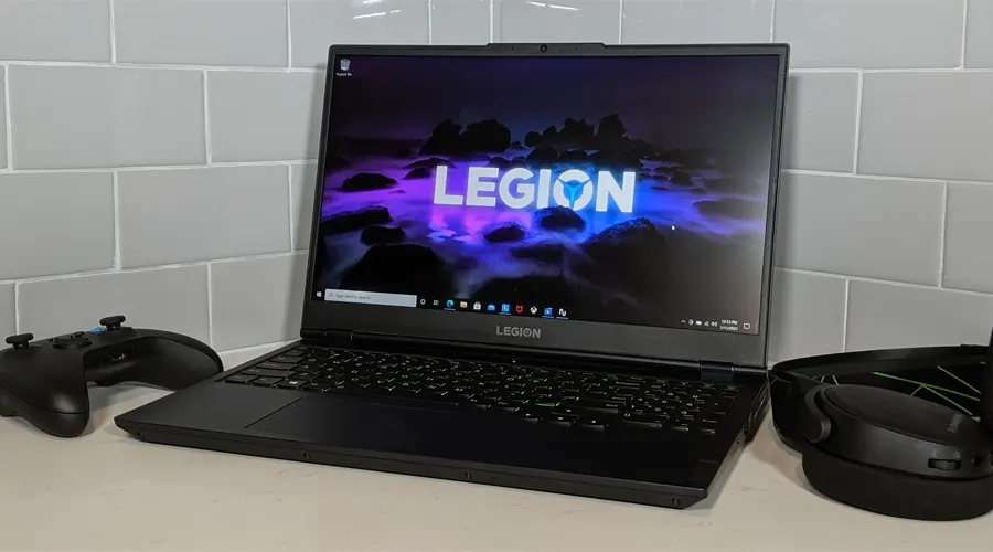 لپ تاپ لنوو مدل Legion 5 روی میز سفید رنگ کنار دیوار طوسی در کنار دسته گیم و هدفون